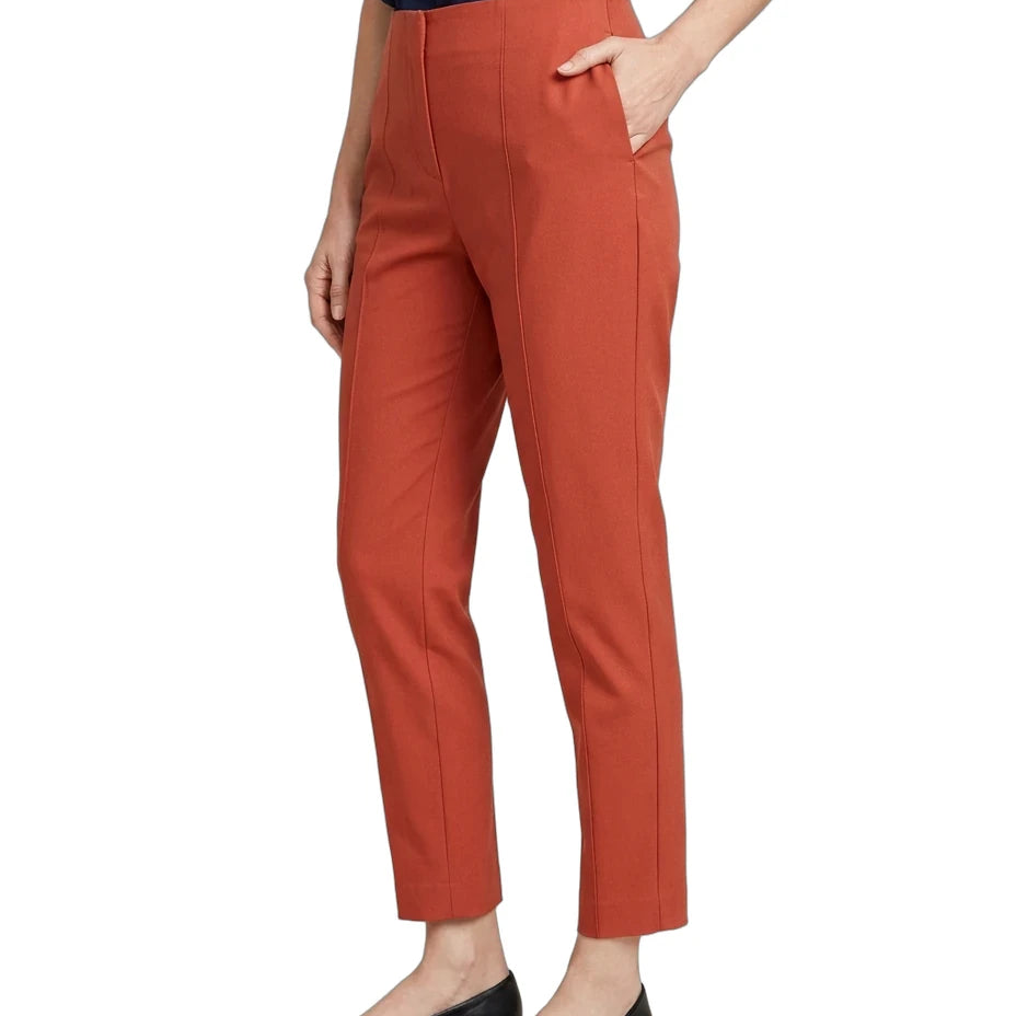 A model wearing a pair of cropped, straight-leg trousers in a warm rust orange color, paired with a navy top and black flats, shown from the waist down on a white background.