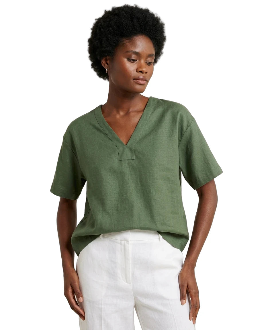 A woman wearing a short-sleeve olive green V-neck top and white pants, photographed against a plain white studio background.
