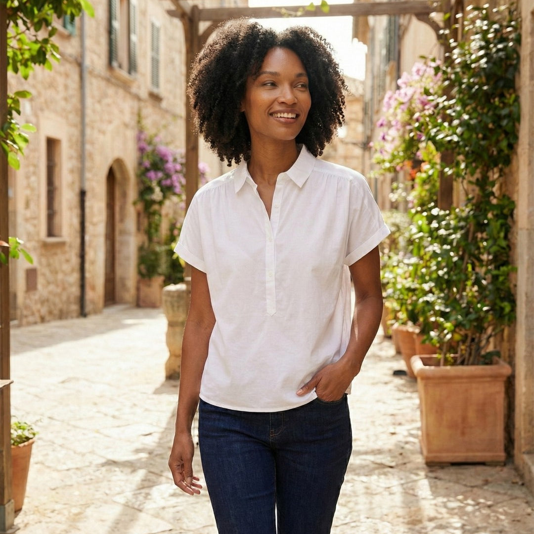 Woman in a white shirt and dark pants standing in a sunlit outdoor setting with plants and stone buildings.