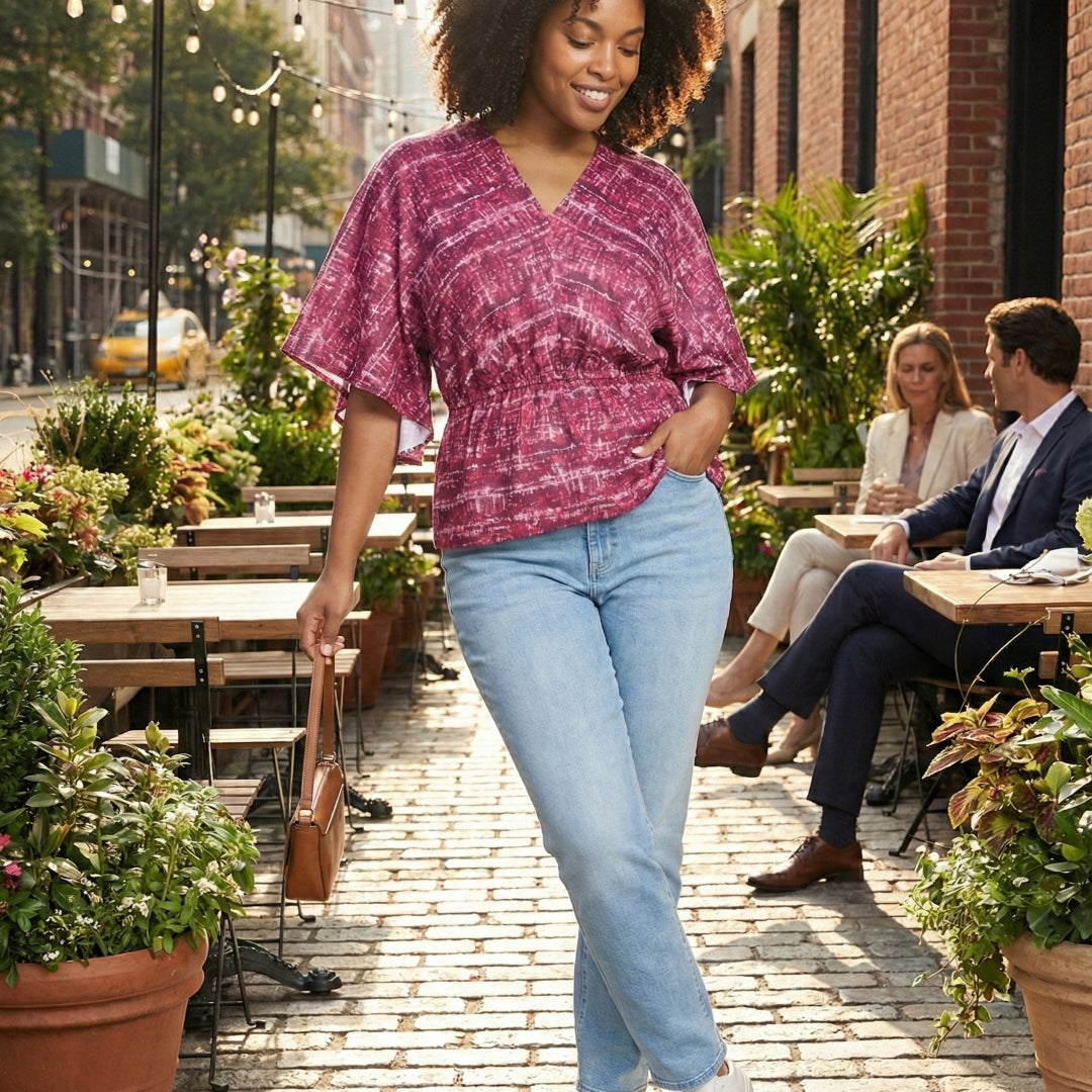 Woman in a pink top and light blue jeans walking on a sidewalk with outdoor seating in the background.