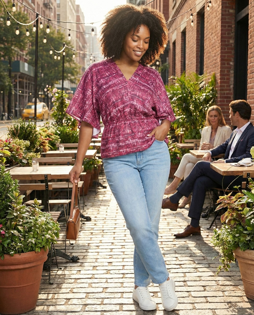 Woman in a pink top and light blue jeans walking on a sidewalk with outdoor seating in the background.