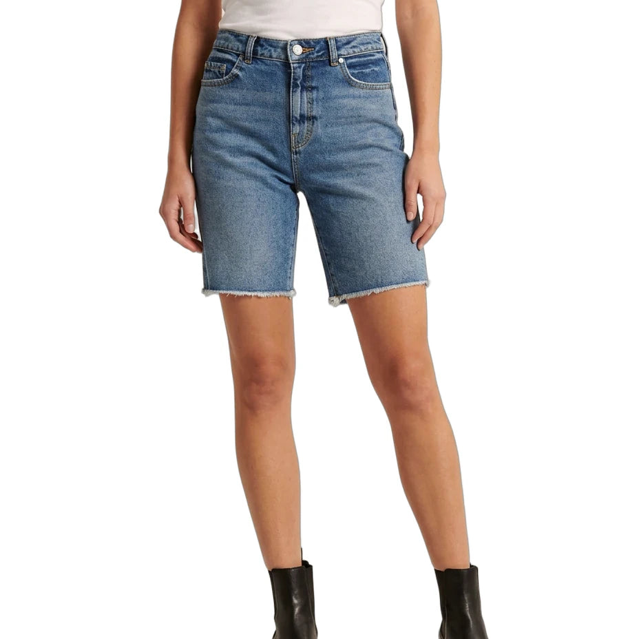 A model wearing medium-wash blue denim shorts with a raw, frayed hem, paired with a white top and black ankle boots, shown against a plain white background.