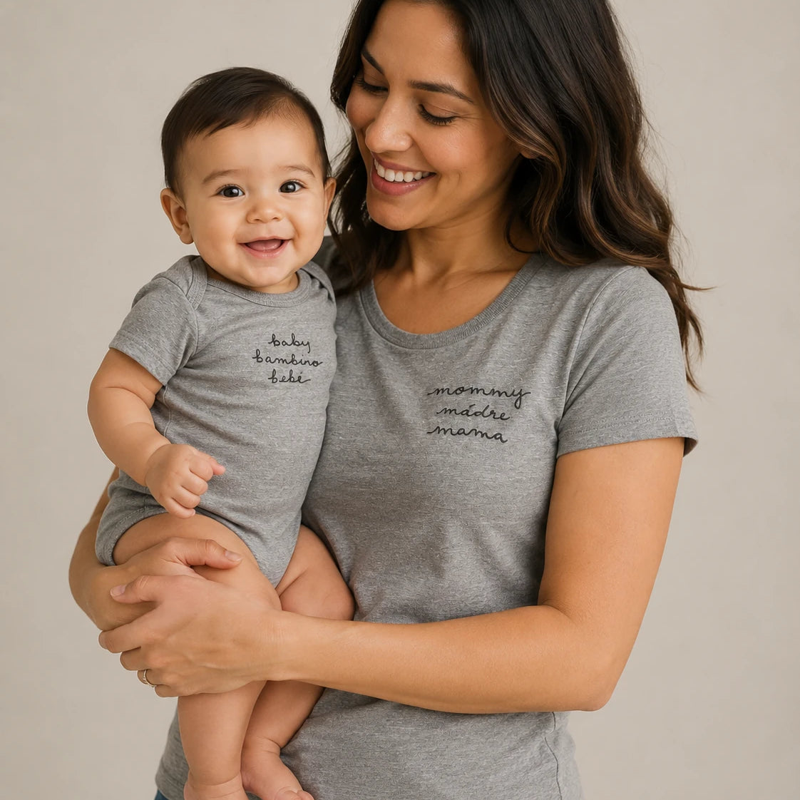 A woman holding a baby, both wearing matching heather gray t-shirts with black script text on the chest, photographed against a plain light beige studio background.
