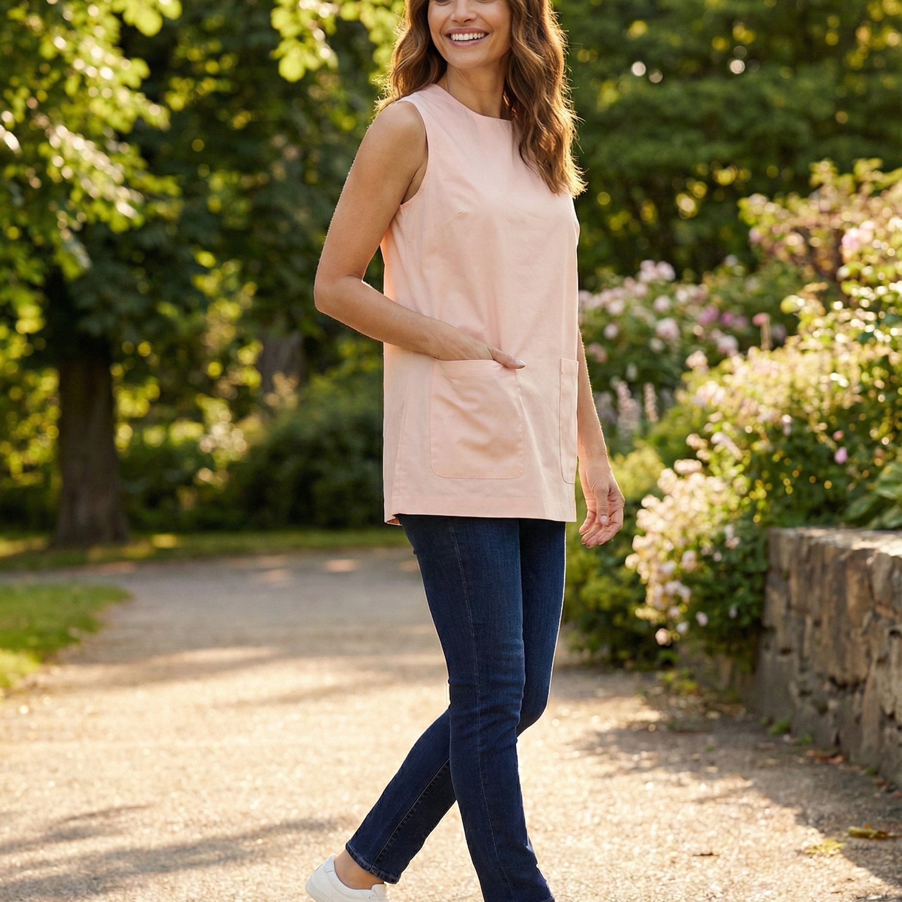 Woman walking outdoors in a park wearing a light pink sleeveless top and dark blue jeans.