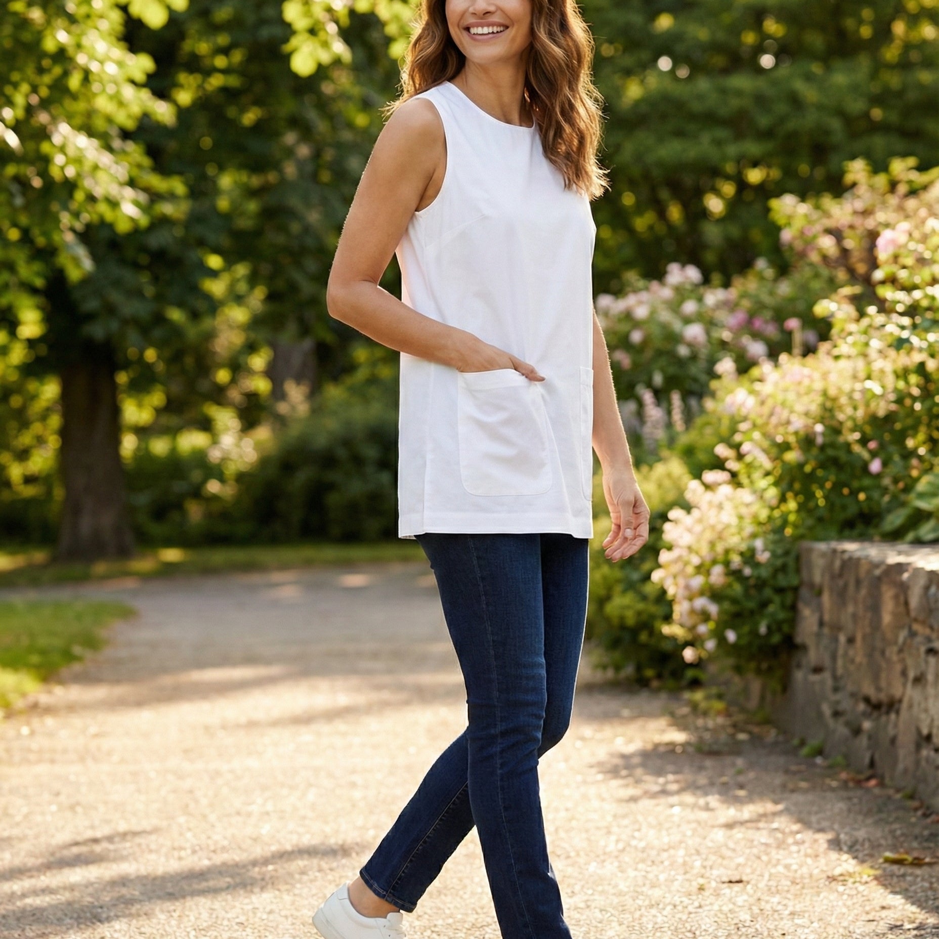 Woman in a white sleeveless top and blue jeans walking outdoors in a garden.