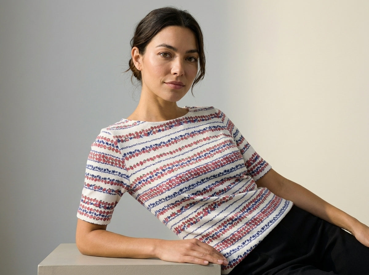 A woman wearing a short-sleeve knit top with horizontal red, blue, and white textured stripes, leaning on a light gray block against a neutral studio background.