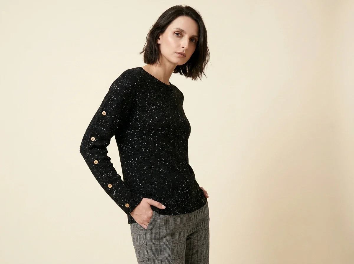 Brunette woman modeling a black speckled knit crewneck sweater with button accents down the sleeves, paired with grey plaid trousers.