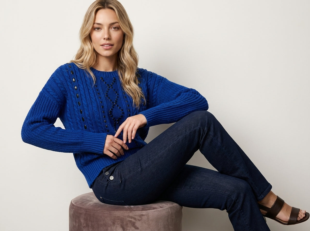 A woman sits on a round velvet stool wearing a textured royal blue cable-knit sweater and dark blue jeans against a plain light gray background.