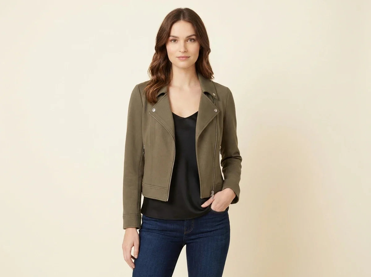 A woman modeling a cropped olive green moto jacket over a black top and blue jeans, standing against a plain beige studio background.