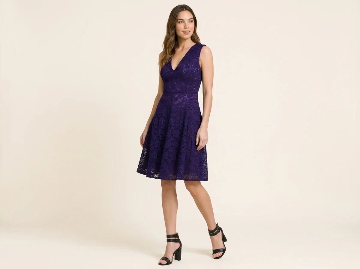 A woman modeling a sleeveless, V-neck, knee-length purple lace dress with a flared skirt, paired with black strappy high-heeled sandals, against a solid light beige studio background.