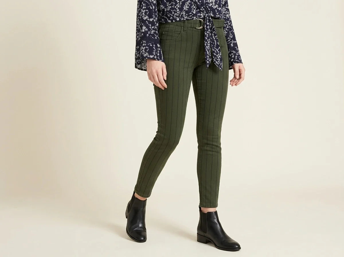 A model wearing olive green pinstriped skinny pants, styled with a dark patterned shirt and black ankle boots, against a plain beige studio background.