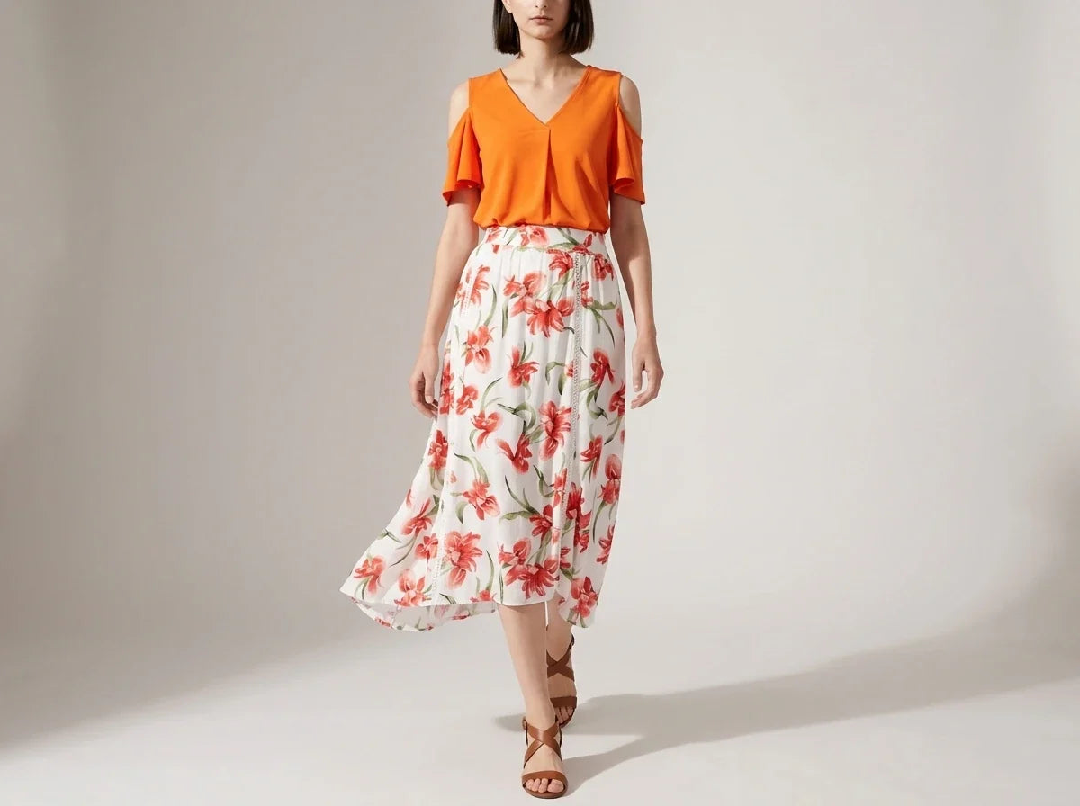 A woman modeling a two-piece outfit consisting of a bright orange cold-shoulder V-neck top and a white midi skirt with a red and green floral print, set against a plain light gray studio background.