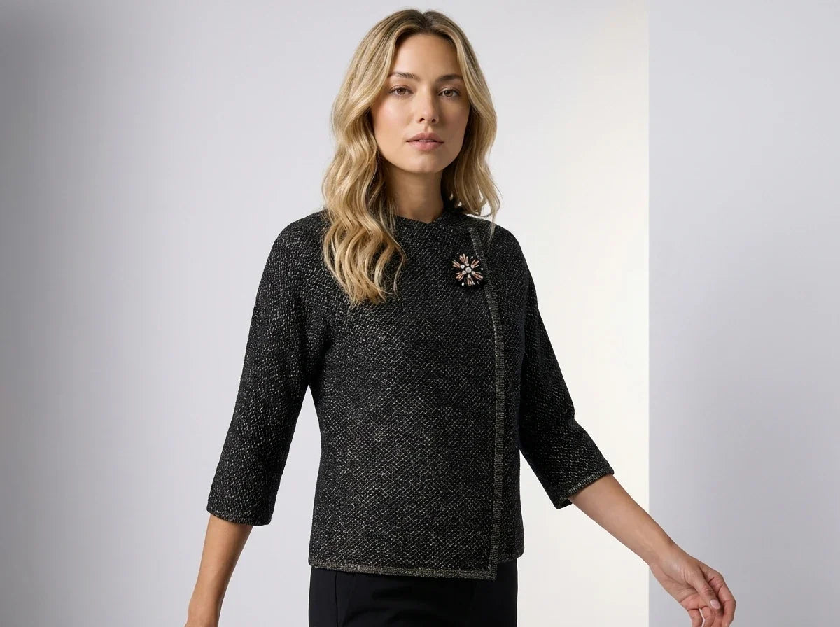 A woman modeling a black textured tweed jacket with three-quarter sleeves, a high mock neck, and a decorative brooch on the chest, set against a neutral studio background.