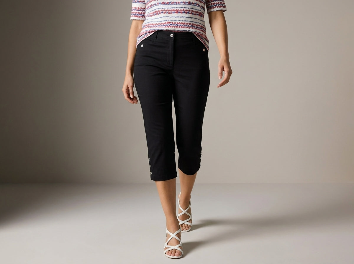 A model wearing black cropped capri pants, a striped short-sleeve top, and white strappy high-heeled sandals, walking against a neutral studio background.