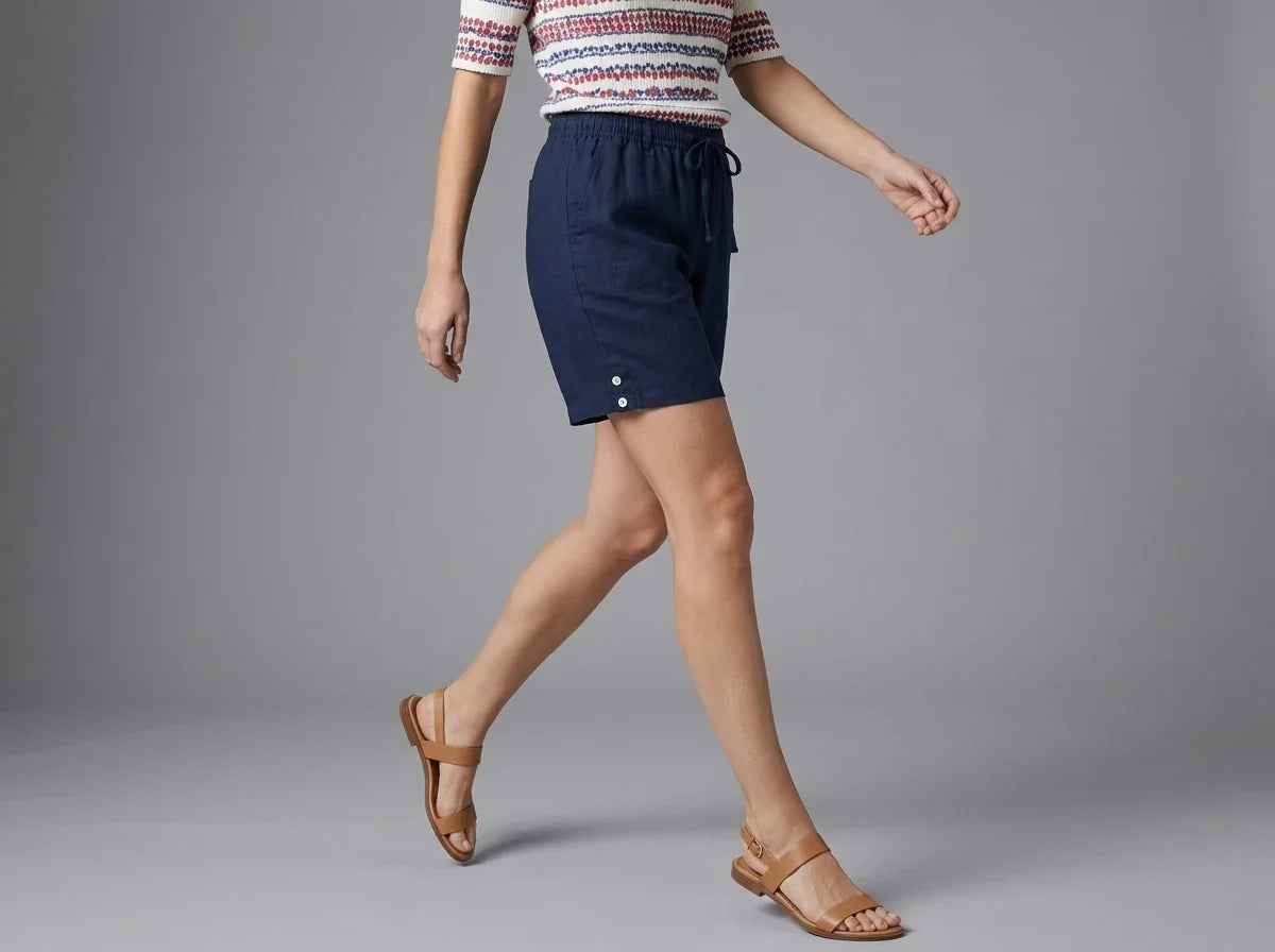 Cropped view of a woman wearing navy blue drawstring shorts with side-hem button details and tan leather strap sandals against a grey studio backdrop.