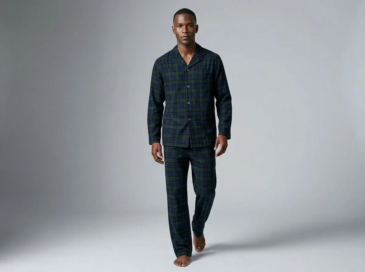 A man modeling a two-piece long-sleeve pajama set with a dark green and black plaid pattern, walking against a neutral gray studio background.