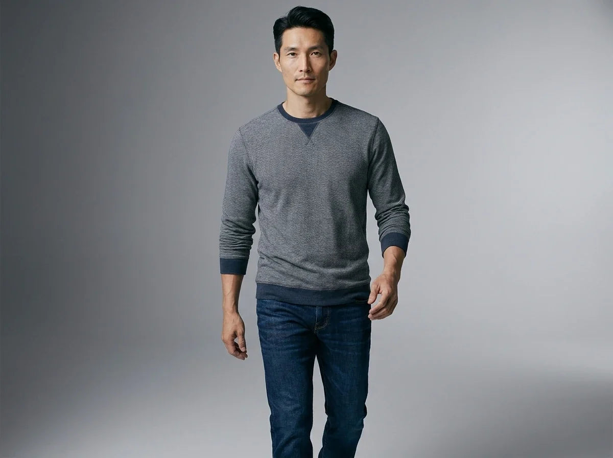 A man wearing a gray long-sleeve crewneck sweater with dark navy trim and blue jeans, standing against a neutral gray studio background.