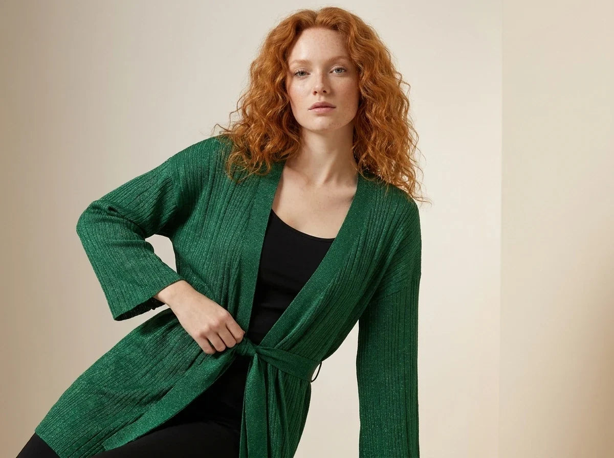 Red-haired woman modeling an emerald green ribbed knit belted cardigan sweater over a black top in a studio setting.
