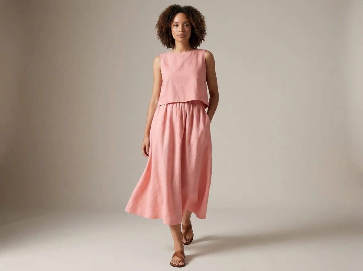 Woman with curly hair modeling a blush pink sleeveless two-piece linen midi dress and brown flat sandals in a studio setting.