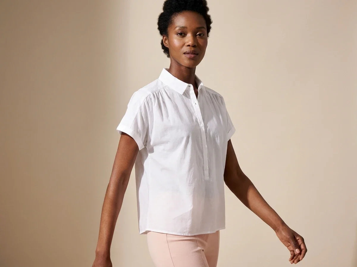 Black woman modeling a white short-sleeve collared button-up blouse with shoulder gathers and light pink pants in a studio setting.