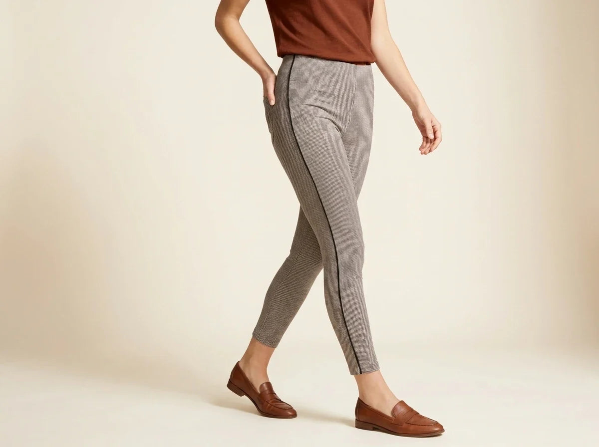 A model wearing light gray heathered leggings with a thin black side stripe, paired with a brown sleeveless top and brown leather loafers, shown from the waist down against a warm beige studio background.