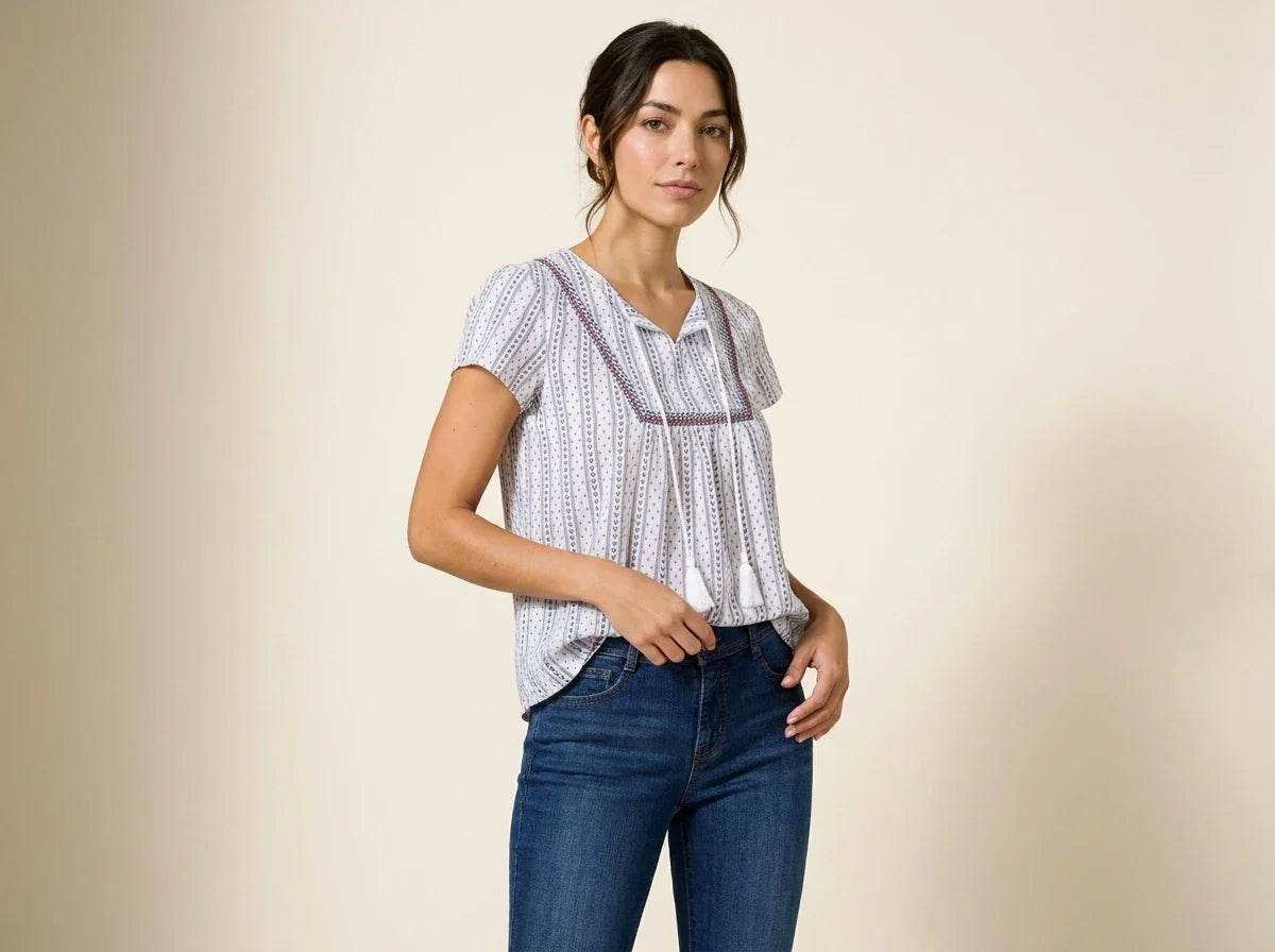 Brunette woman modeling a white short-sleeve peasant blouse with vertical blue dotted stripes and embroidered tassel ties, paired with dark blue jeans.