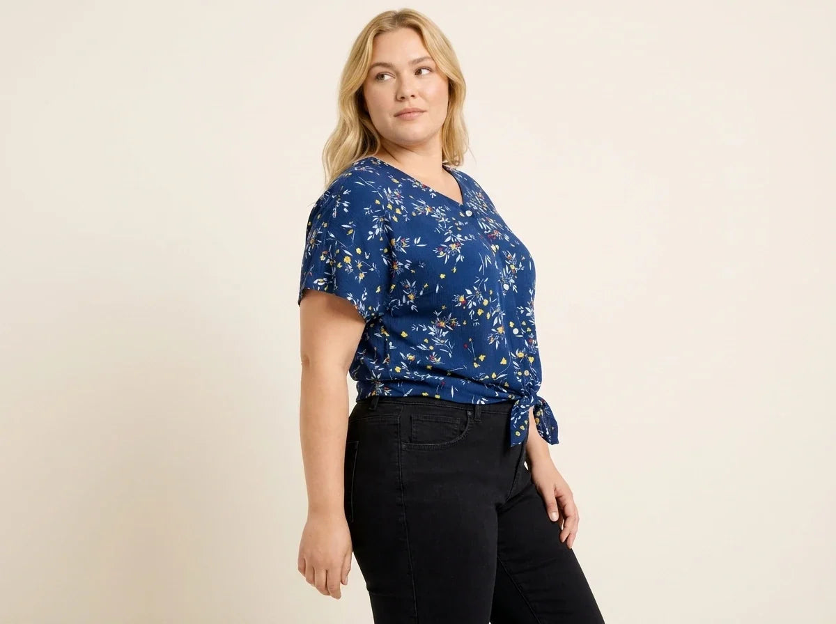 Blonde woman modeling a plus-size cobalt blue floral short-sleeve top with a side-tie knot hem paired with black jeans.