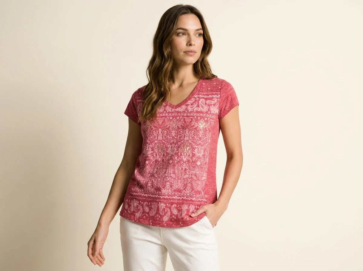 Brunette woman modeling a raspberry pink short-sleeve V-neck t-shirt with a paisley tapestry graphic print paired with white pants.