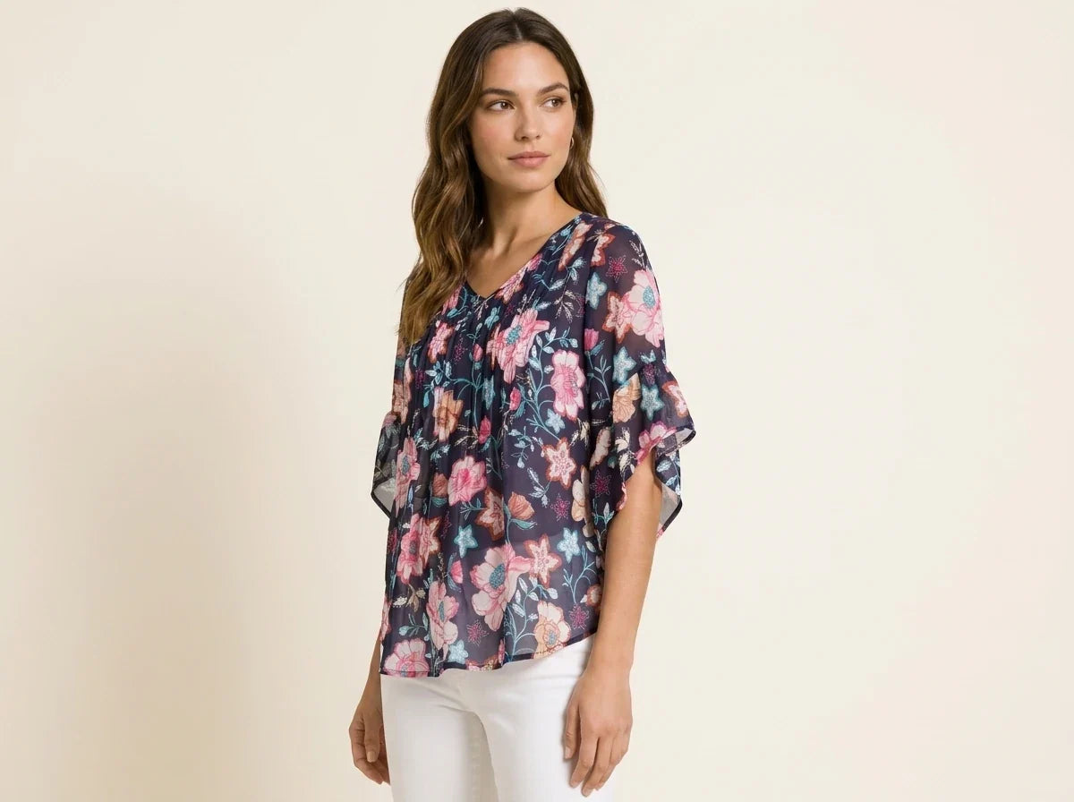 Brunette woman modeling a navy blue floral print chiffon blouse with tiered flutter sleeves paired with white pants.