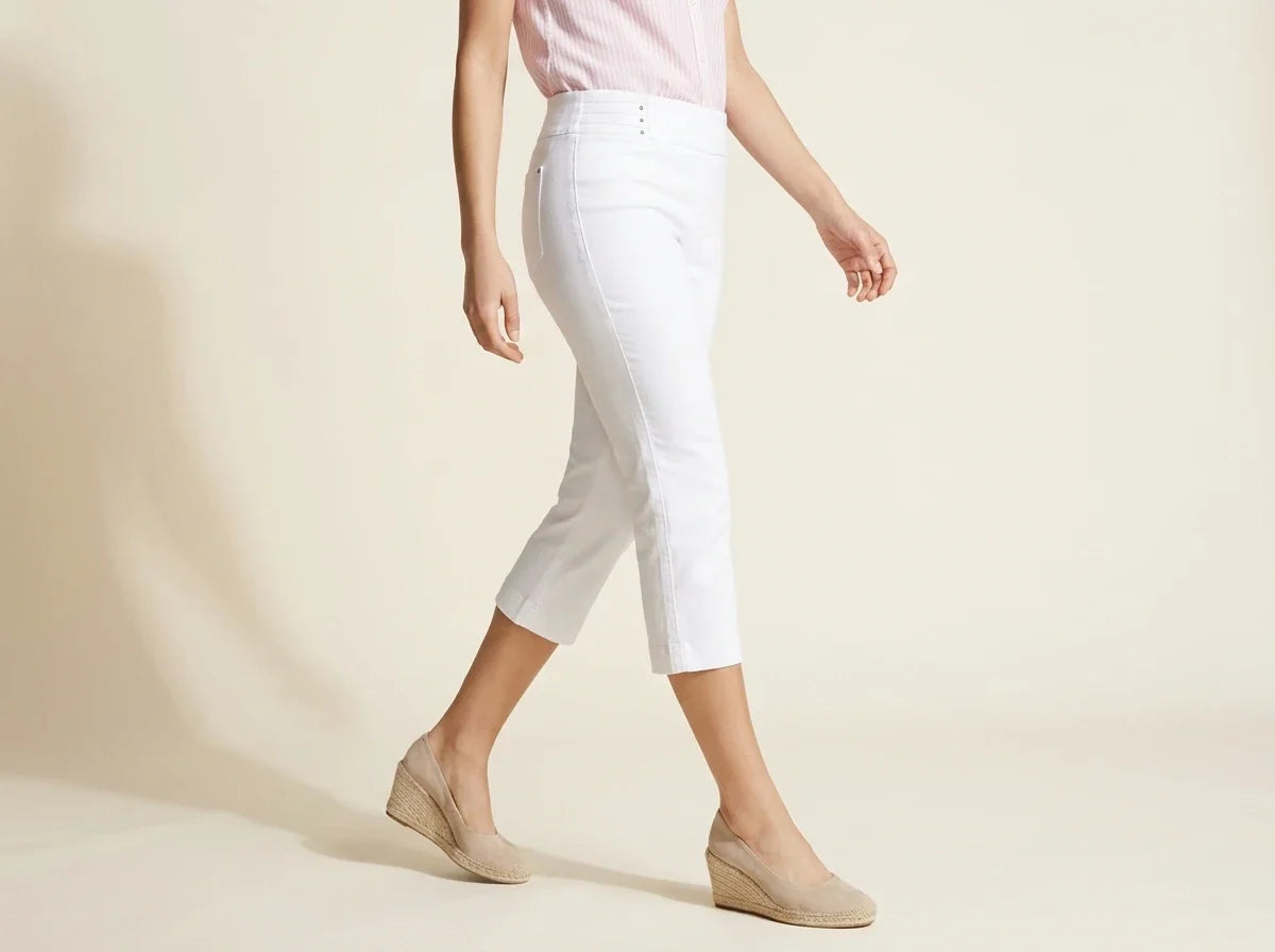 A model wearing white cropped capri pants, a light pink top, and beige wedge espadrilles, walking against a warm beige studio background.