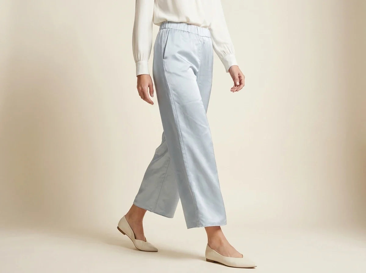 A model wearing light blue wide-leg cropped trousers, a white long-sleeve top, and beige flats, shown from the waist down against a warm beige studio background.