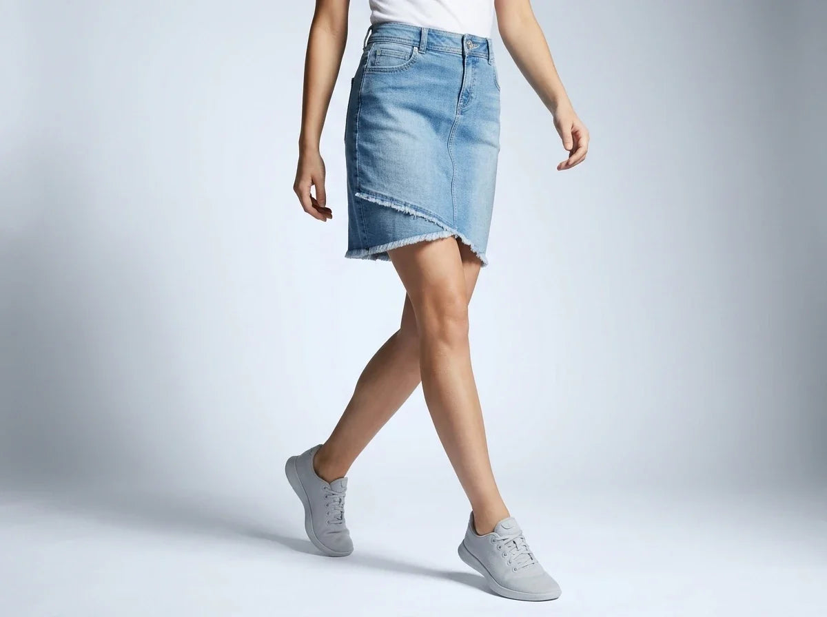 A model wearing a light blue denim asymmetrical mini skirt with frayed edges, paired with a white top and gray sneakers, captured against a light gray studio background.