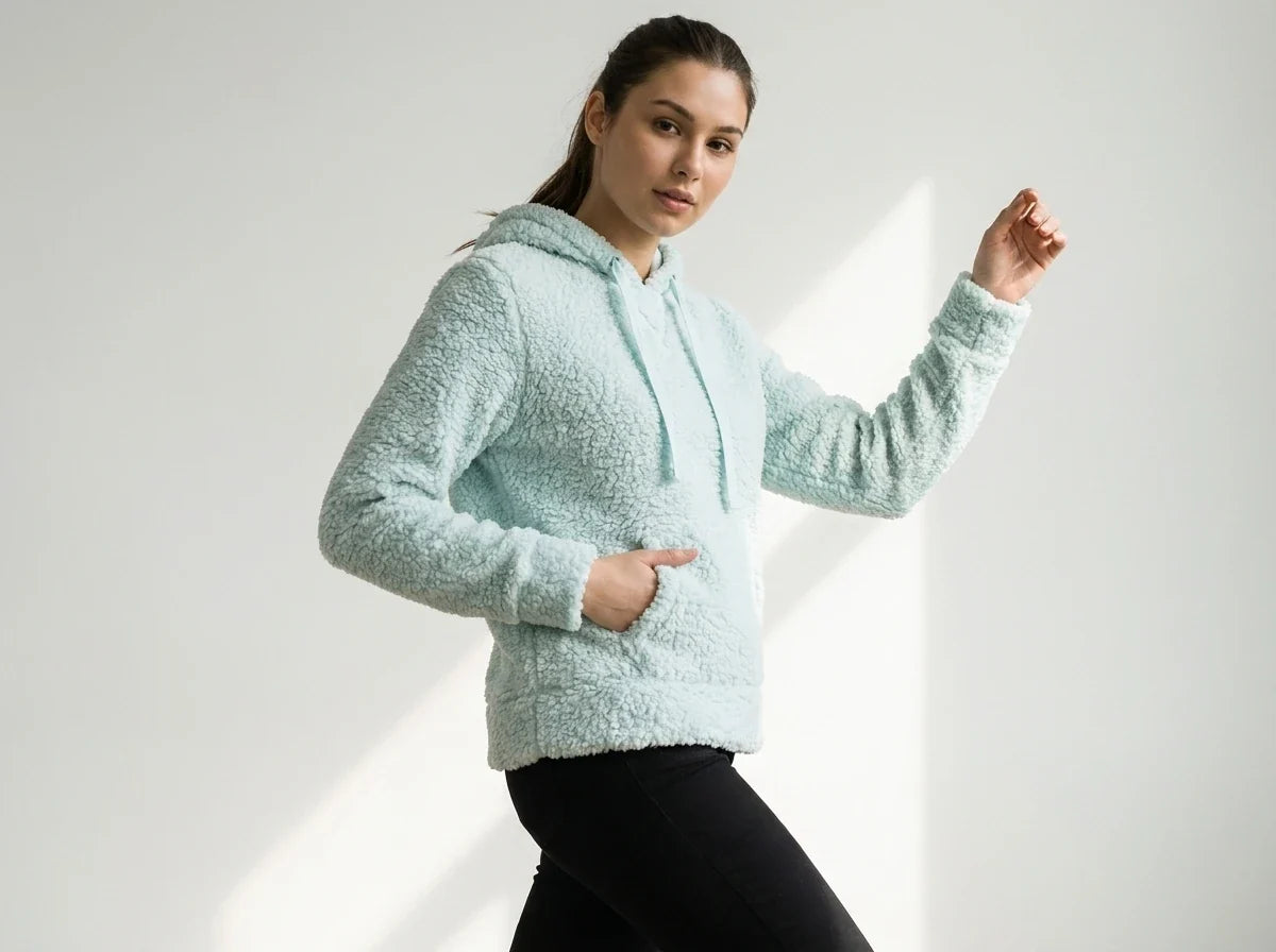 A woman modeling a light mint green sherpa fleece hoodie with black pants, posing against a white studio background with soft diagonal shadows.
