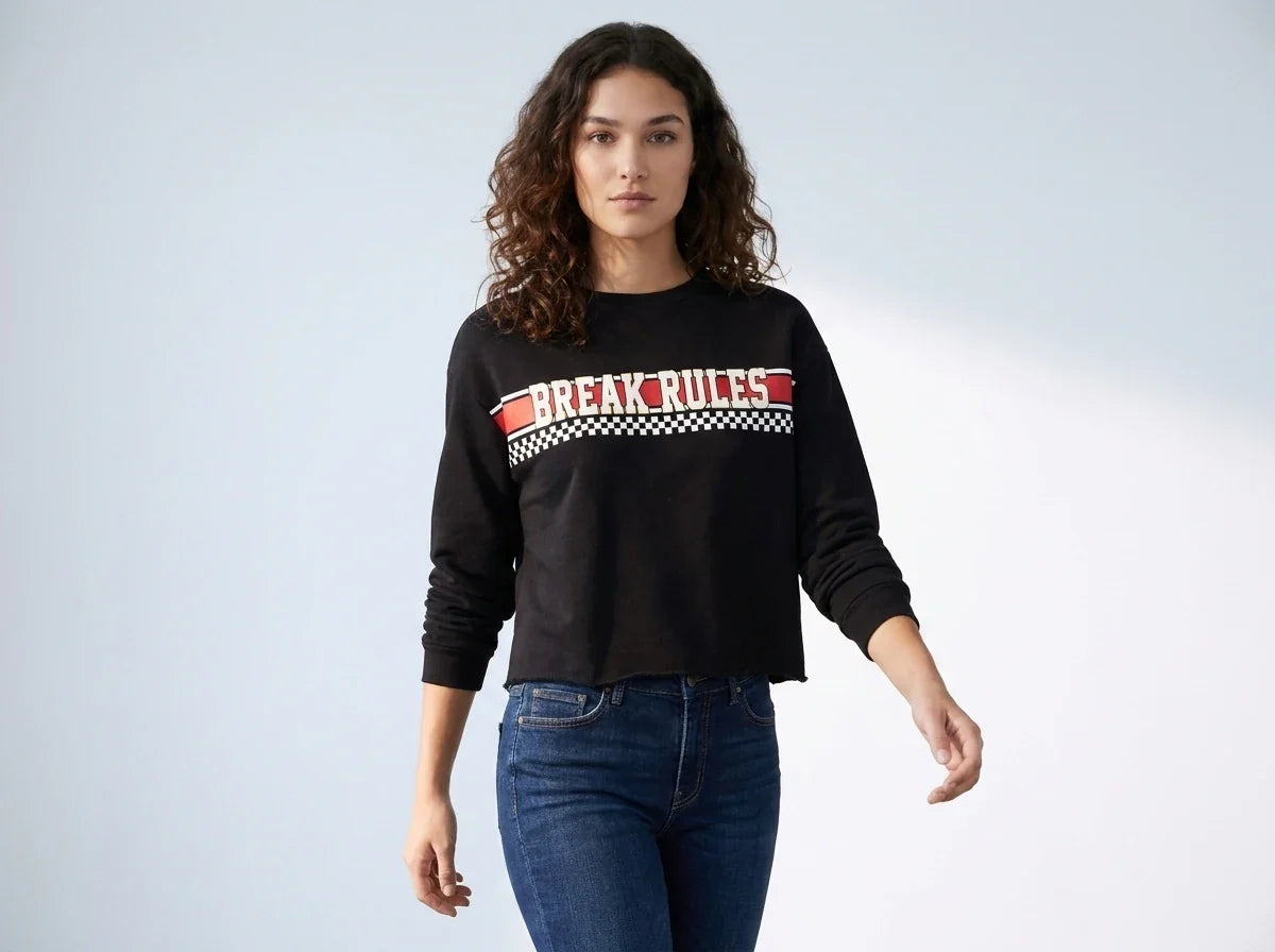 A woman modeling a black long-sleeve cropped sweatshirt with a red and white 'BREAK RULES' graphic across the chest, paired with blue jeans, against a light gray studio background.