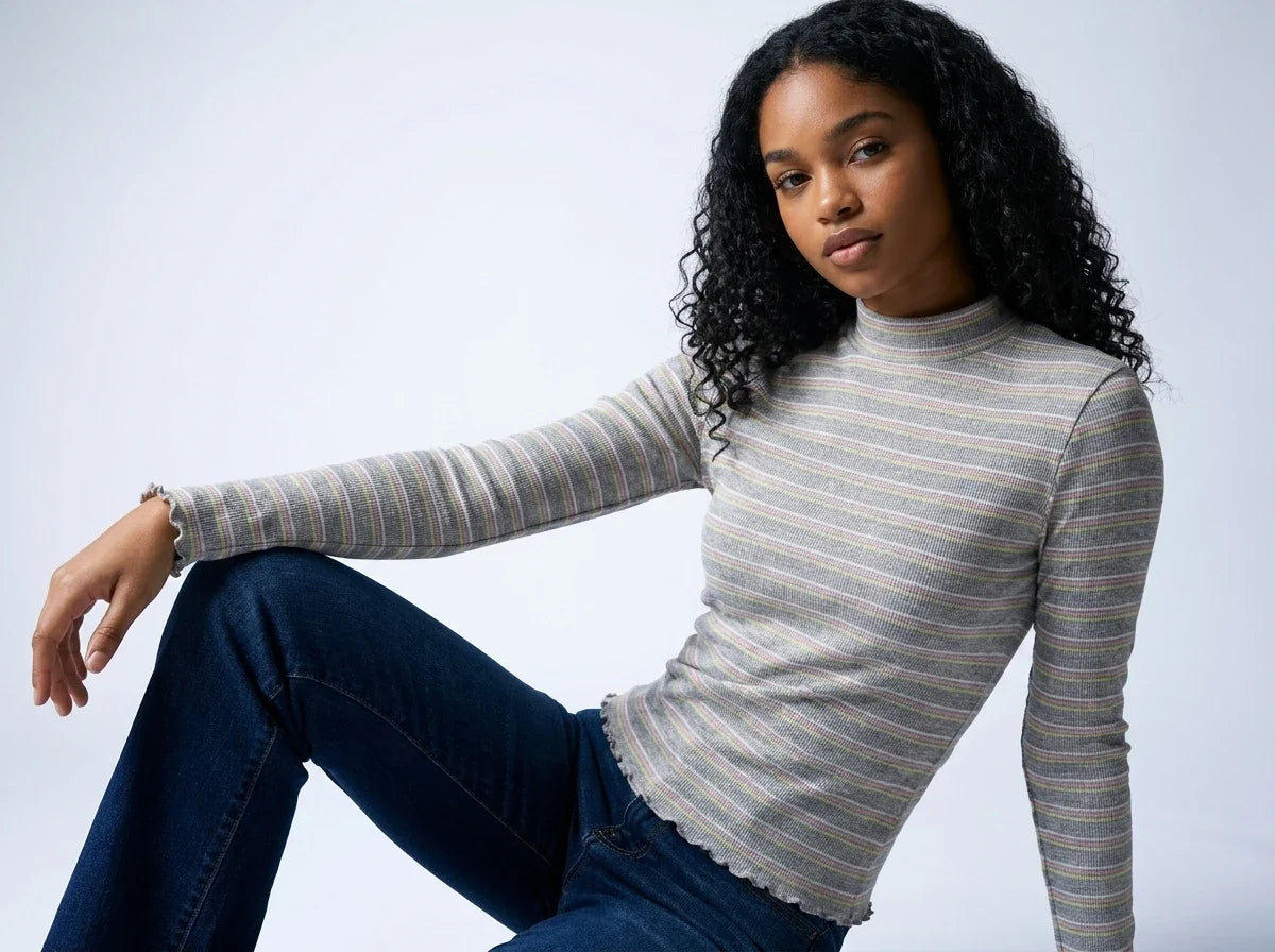 A woman with curly black hair wearing a gray and white striped mock neck long-sleeve top and dark blue jeans, posing against a light gray studio background.