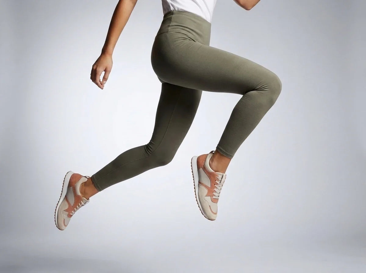 Side view of a woman mid-air wearing olive green high-waisted leggings and neutral athletic sneakers.