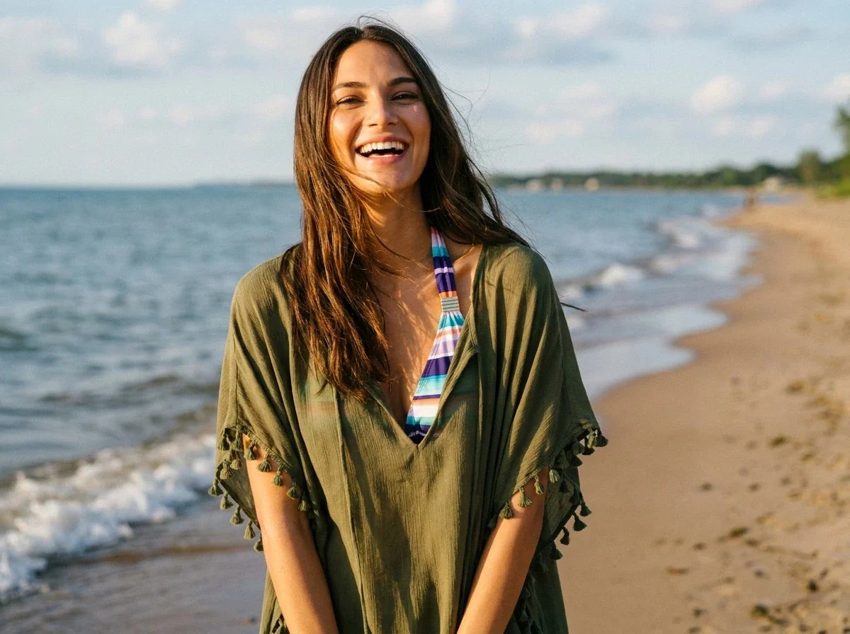 A woman smiling on a beach wearing an olive green bohemian tunic cover-up over a colorful striped bikini top, with the ocean and sandy shore in the background.
