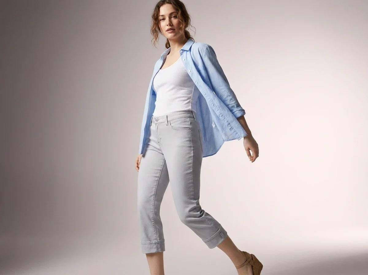 A woman modeling a light blue button-up shirt over a white tank top and light gray cropped jeans, standing against a neutral studio background.