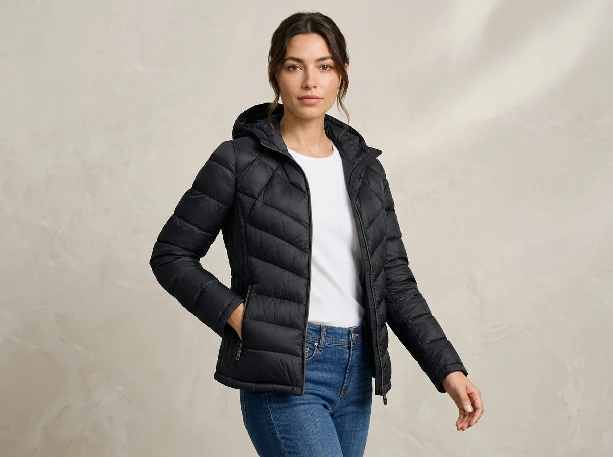 A woman wearing a black hooded puffer jacket over a white top and blue jeans, standing against a textured light beige studio backdrop.