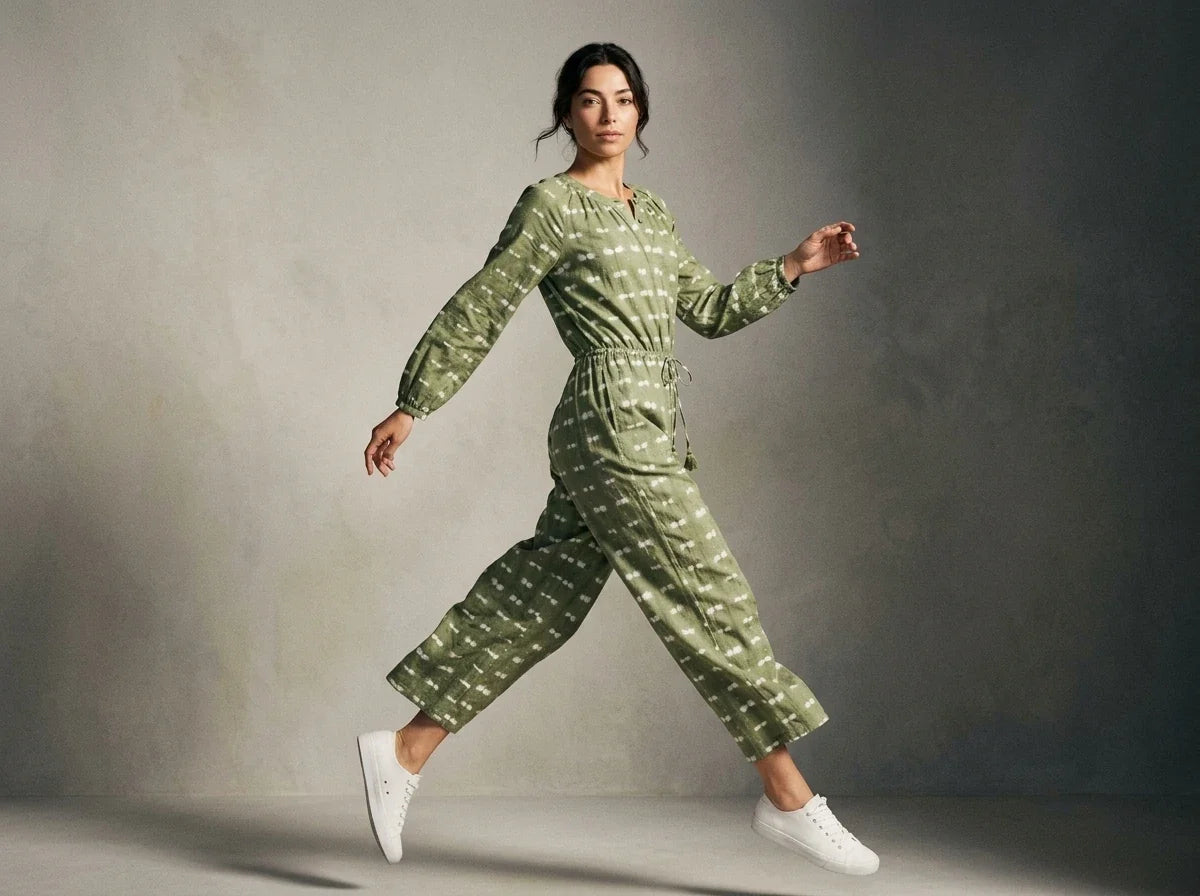 A woman wearing a sage green long-sleeve jumpsuit with a white geometric pattern and white sneakers, captured in a dynamic pose against a textured gray studio backdrop.