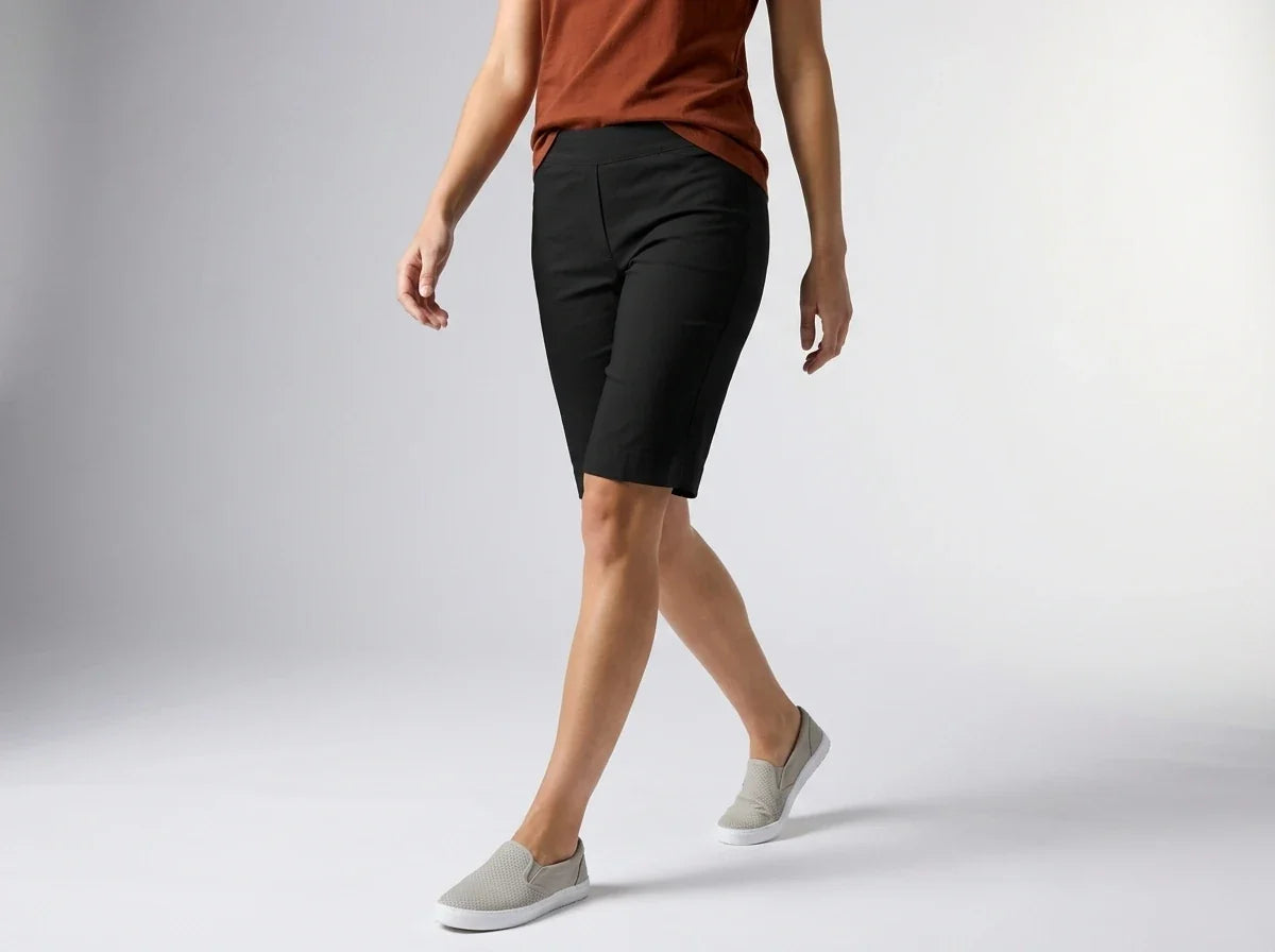 A model wearing black knee-length athletic shorts, a rust-orange top, and light gray slip-on sneakers, captured from the waist down against a light gray studio background.