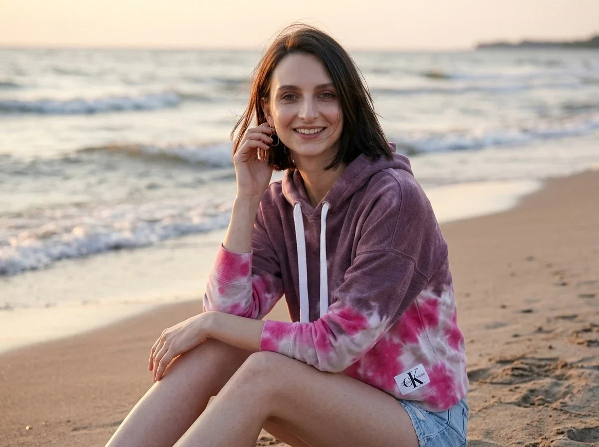 A woman sitting on a sandy beach at sunset wearing a pink and purple tie-dye hoodie with white drawstrings and a small Calvin Klein logo patch near the hem.