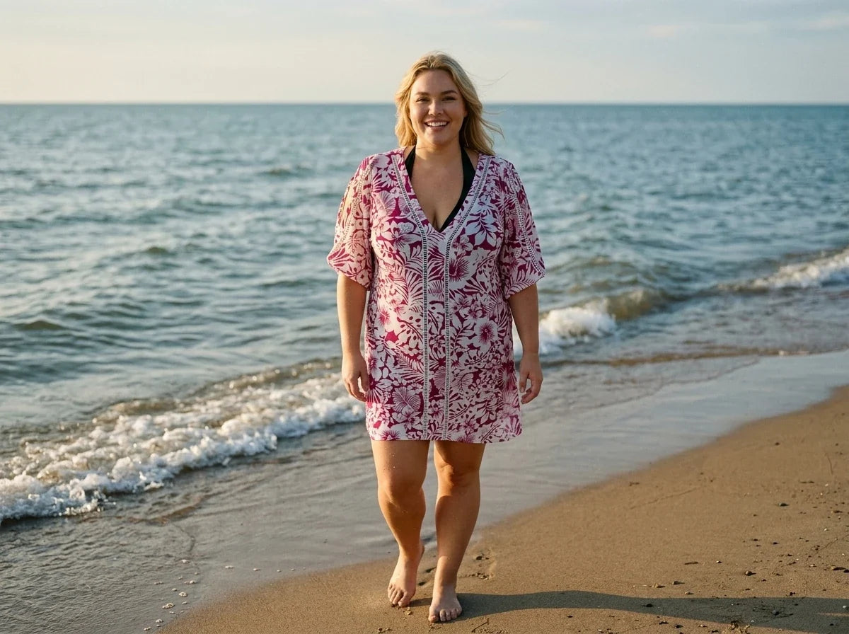 A woman walking on a sandy beach wearing a pink and white floral print short-sleeve cover-up with black trim over a black swimsuit.