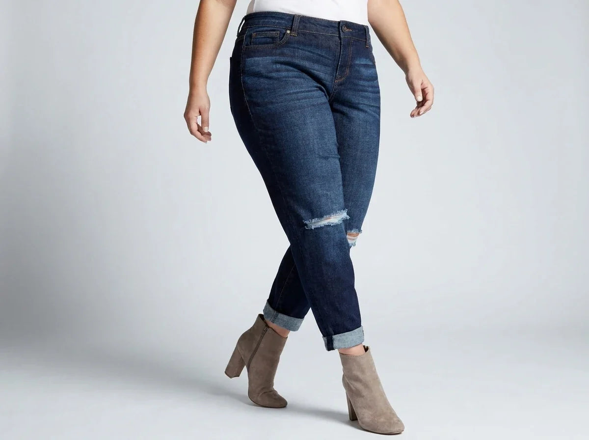 A model wearing dark blue distressed skinny jeans with cuffed hems, paired with a white shirt and tan ankle boots, standing against a light gray studio background.