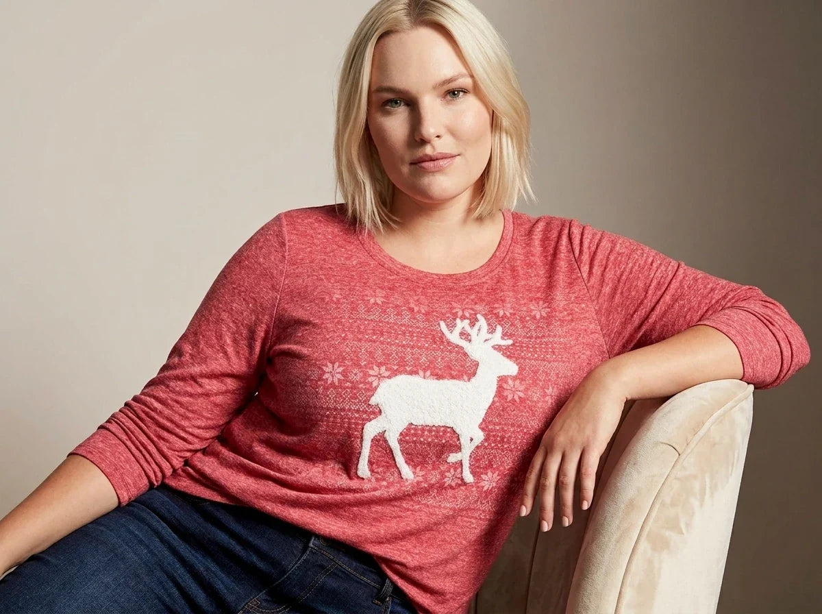 A woman wearing a heathered red long-sleeve knit sweater with a white reindeer and snowflake pattern, paired with dark blue jeans, sitting on a beige armchair against a neutral studio background.