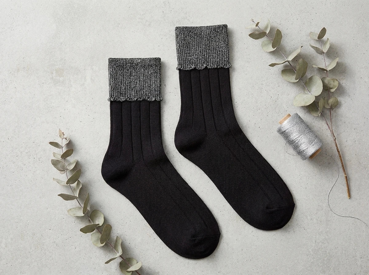 "Flat lay of women's black ribbed crew socks with metallic silver folded cuffs, styled with eucalyptus branches and silver thread spool.
