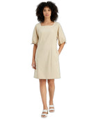 A woman modeling a light beige midi dress with puff sleeves, a square neckline, and side pockets, paired with white strappy sandals against a plain white background.