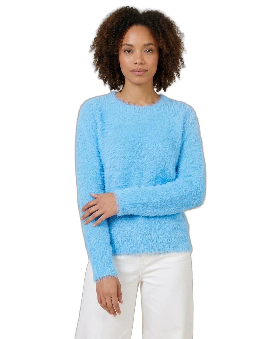 A woman with curly brown hair wearing a fuzzy light blue long-sleeve sweater and white pants, standing against a plain white background.