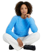 A woman with curly dark hair sits cross-legged against a white background, wearing a fuzzy bright blue long-sleeve sweater, white pants, and black boots.