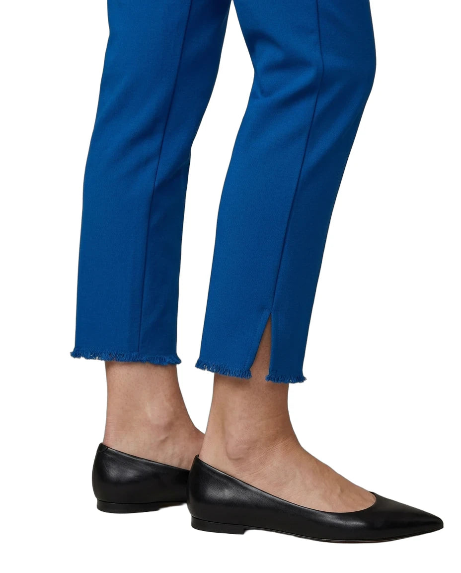 A close-up of a person's legs wearing bright blue cropped trousers with a raw frayed hem and side slits, paired with black pointed-toe flats, against a plain white background.